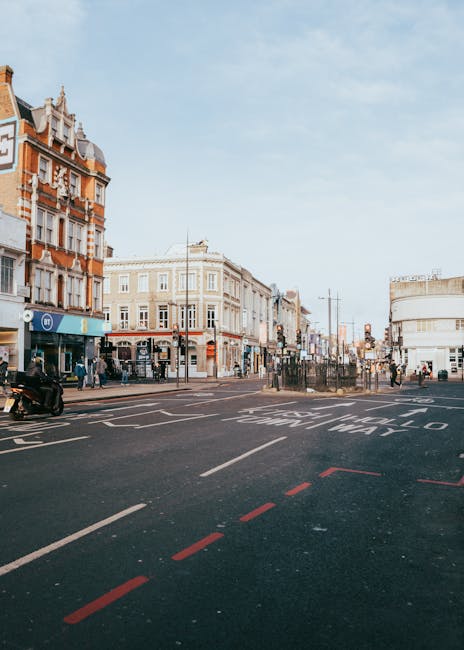 A wide view of a busy street in Ealing, with classic Victorian-style buildings featuring ornate facades and decorative elements on the left side, and modern commercial buildings, including a curved white structure, on the right. The street has marked lanes and red dashed lines indicating a parking or loading zone, with a few parked motorcycles and bicycles visible. Pedestrians are walking along the pavement, some crossing the street at designated crosswalks. The sky is partly cloudy with a soft daylight illuminating the scene, typical of urban activity related to house and office removals. This setting exemplifies the typical environment where Ealing Man and Van conducts relocation logistics, including furniture transport and packing services, in the context of home or business moves.