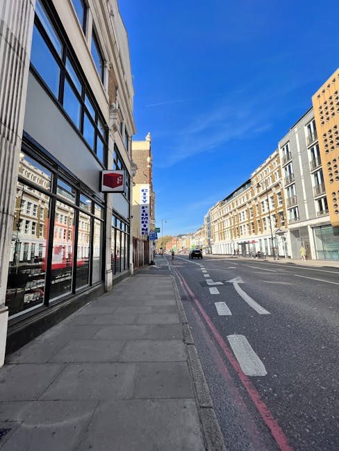 A clear daytime street scene outside a commercial building with large glass windows displaying merchandise. The pavement runs alongside the building, with a few small stacks of cardboard boxes and wrapped furniture items placed near the entrance, indicating a house or office removal process. Visible moving blankets, plastic wrapping, and packing materials are used to protect furniture on the pavement, with some items being lifted or carried by movers from the building toward a waiting van. A loading area is set up with a vehicle partially visible further down the street, suggesting ongoing furniture transport as part of a professional removals service. The street features parked cars and building facades consistent with an urban area, supporting the context of a local home or business relocation, such as those offered by Ealing Man and Van, operating in Ealing and surrounding areas.