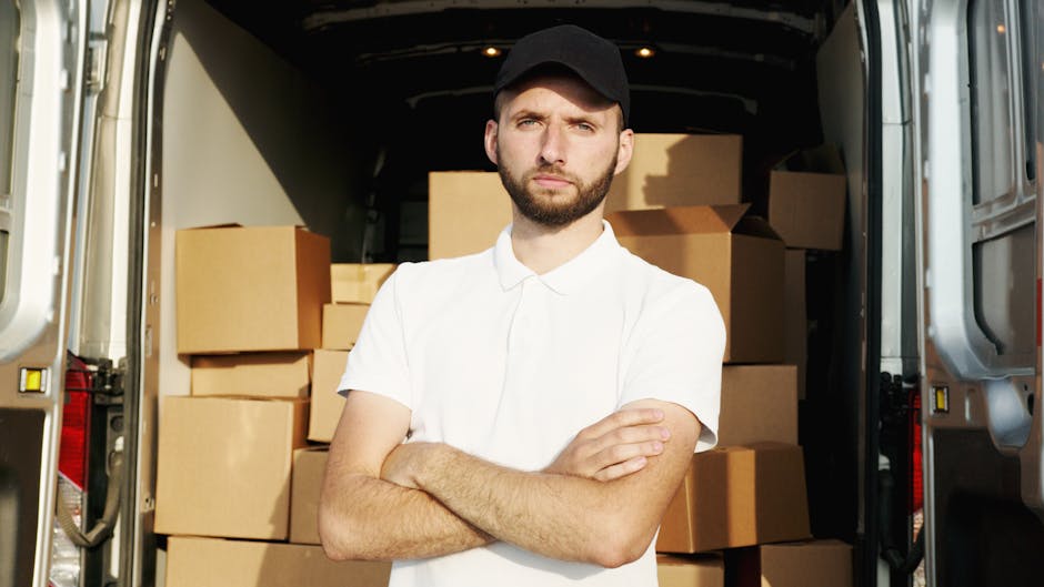 A young man with a beard, wearing a black cap and white polo shirt, stands inside the open rear of a moving van loaded with multiple cardboard boxes of various sizes. The boxes are stacked neatly on the vehicle's floor, some wrapped in plastic or packing material, indicating they are packed for a home relocation. The van has a metallic interior, and the boxes are positioned to facilitate loading or unloading during moving services. Outside the van, part of the pavement is visible, suggesting the loading area. The man appears to be observing the loading process or preparing to assist with furniture or box transport during a house move, aligned with professional removals service by Ealing Man and Van.