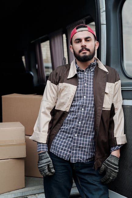 A young man with a beard, wearing a black cap and white polo shirt, stands inside the open rear of a moving van loaded with multiple cardboard boxes of various sizes. The boxes are stacked neatly on the vehicle's floor, some wrapped in plastic or packing material, indicating they are packed for a home relocation. The van has a metallic interior, and the boxes are positioned to facilitate loading or unloading during moving services. Outside the van, part of the pavement is visible, suggesting the loading area. The man appears to be observing the loading process or preparing to assist with furniture or box transport during a house move, aligned with professional removals service by Ealing Man and Van.