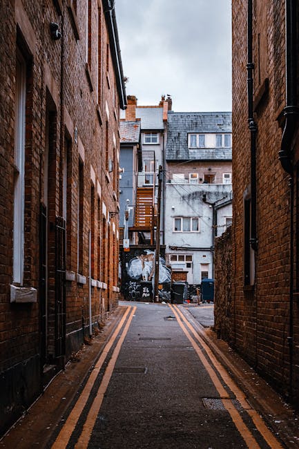 A narrow alleyway between two brick buildings with a paved surface marked by double yellow lines along each side. In the background, there are multiple residential structures, including a grey building with a wooden staircase attached to its exterior. Several black rubbish bins are lined up near the base of the staircase, and a black graffiti-covered wall is visible further along the alley. The surroundings are illuminated by natural daylight, highlighting the urban environment. Ealing Man and Van, a professional removals company, might use such a location for house removals or the transportation of furniture during a home relocation, demonstrating a typical setting for loading or unloading furniture and boxes in challenging street conditions consistent with tight or urban streetscapes.