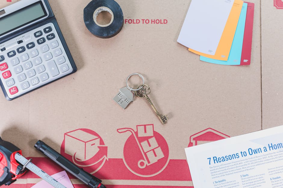 Arrangement of various office and packing supplies on a cardboard surface, including a grey calculator with white and pink buttons, a small roll of black tape, a set of colorful folders or cards (white, orange, blue, red), a metallic keychain with a house-shaped charm, a small black marker, a pink measuring tape, and a printed document titled '7 Reasons to Own a Home'. The background features symbols of moving, such as a box with an arrow, a hand truck with a box, and a house icon, indicating the context of home relocation, packing, and moving services from Ealing Man and Van. The scene depicts preparations for a home removal, emphasizing packing materials, organizational tools, and logistical elements involved in furniture transport and move planning.