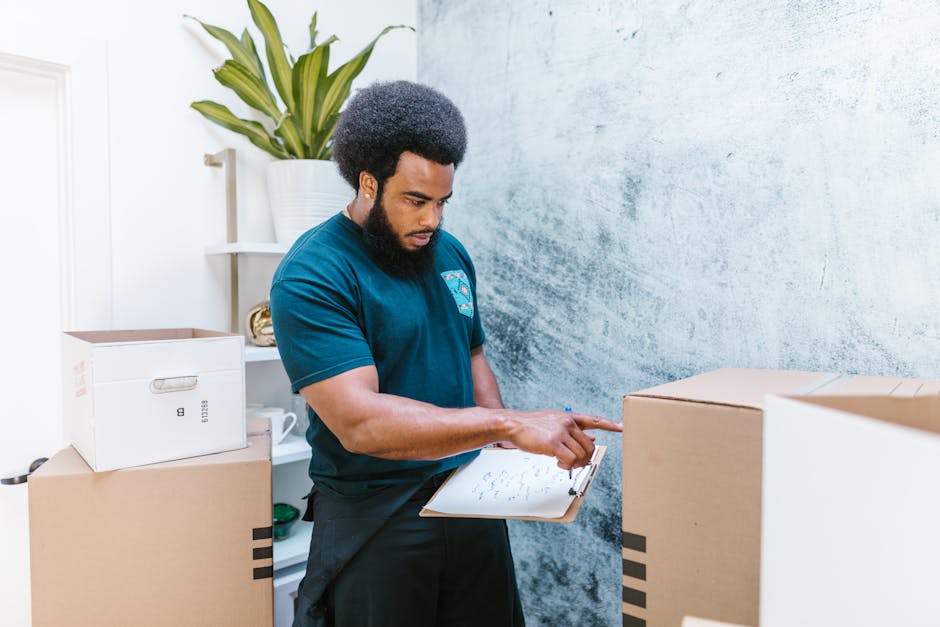 A professional mover from Ealing Man and Van is reviewing a packing inventory clipboard as part of a house removal process inside a residential property. The mover, dressed in a navy blue T-shirt, is standing between several cardboard moving boxes, some sealed and others open, which are placed on a table and the floor. Behind him, there is a white shelving unit holding a potted plant, a ceramic mug, and other small household items. The background features a blue textured wall and natural light illuminating the scene, highlighting the organized packing and loading process typical of furniture transport and home relocation services. The mover appears to be assessing the items carefully, ensuring all belongings are accounted for ahead of transportation, reflecting the detailed planning necessary for effective packing and moving. This visual relates to resource management and logistics involved in house removals, as provided by Ealing Man and Van.